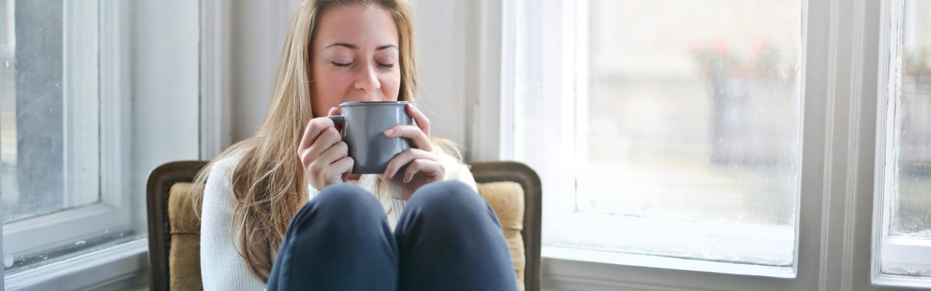 Woman enjoying a warm drink in a cozy armchair by a window, embracing relaxation.